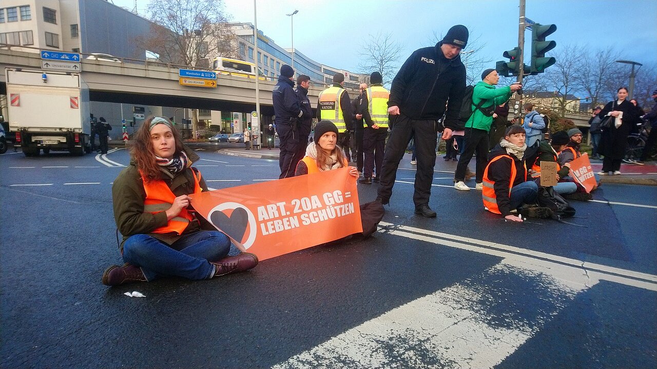 Zwei frauen sitzen mit einem Plakat auf der Straße.Ein Polizist spricht sie an.